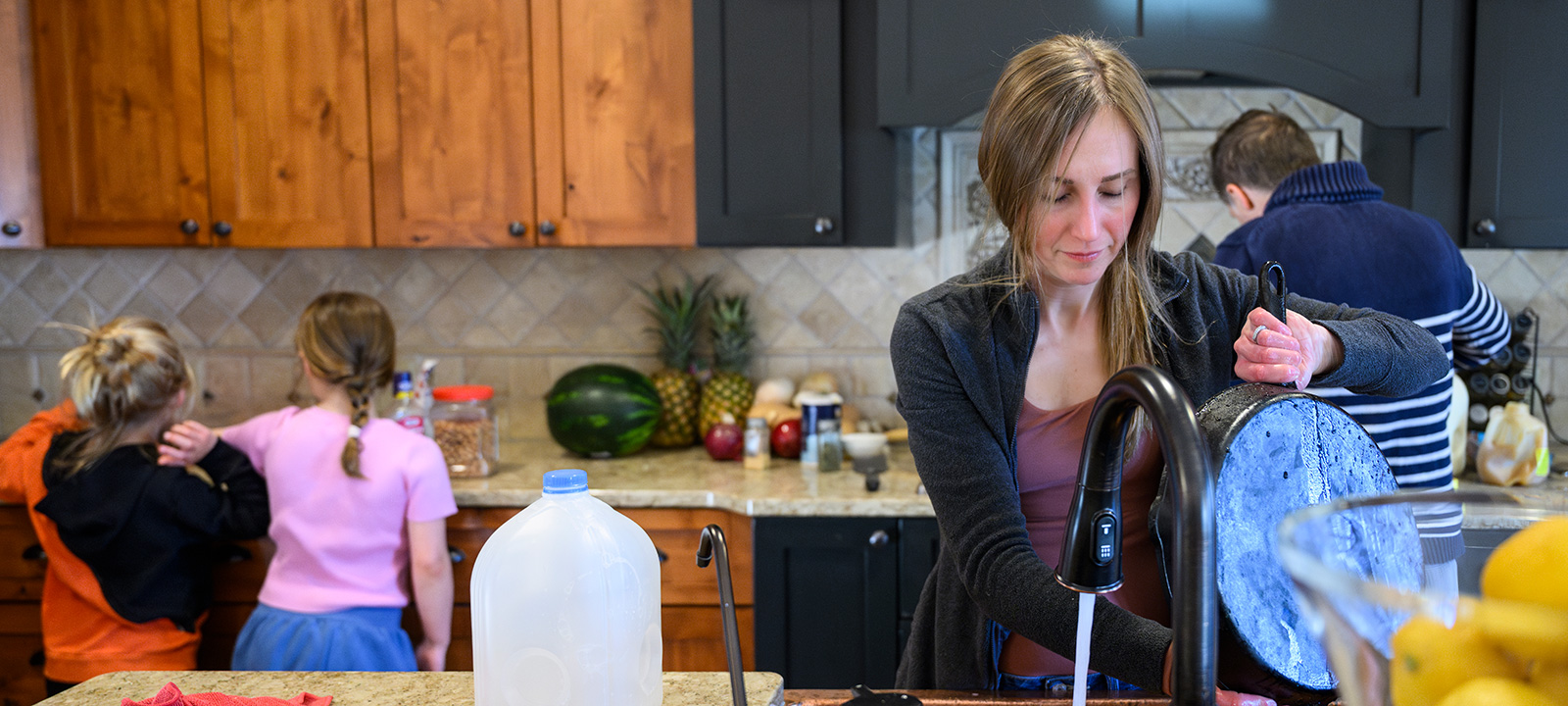 A kitchen scene with Jessica Calarco washing a large pot at the sink, two children standing at the counter, and her husband cooking at the stove, surrounded by items like a gallon of milk, pineapples, watermelon, and kitchen utensils.