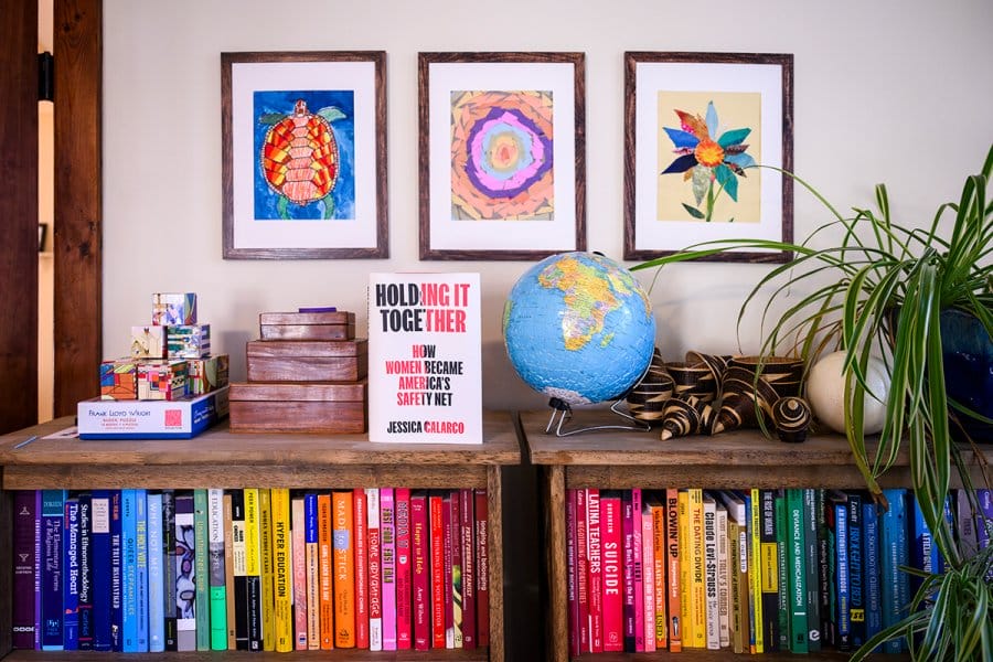 A colorful bookshelf with books arranged by color, a globe, a plant, and framed artwork above.