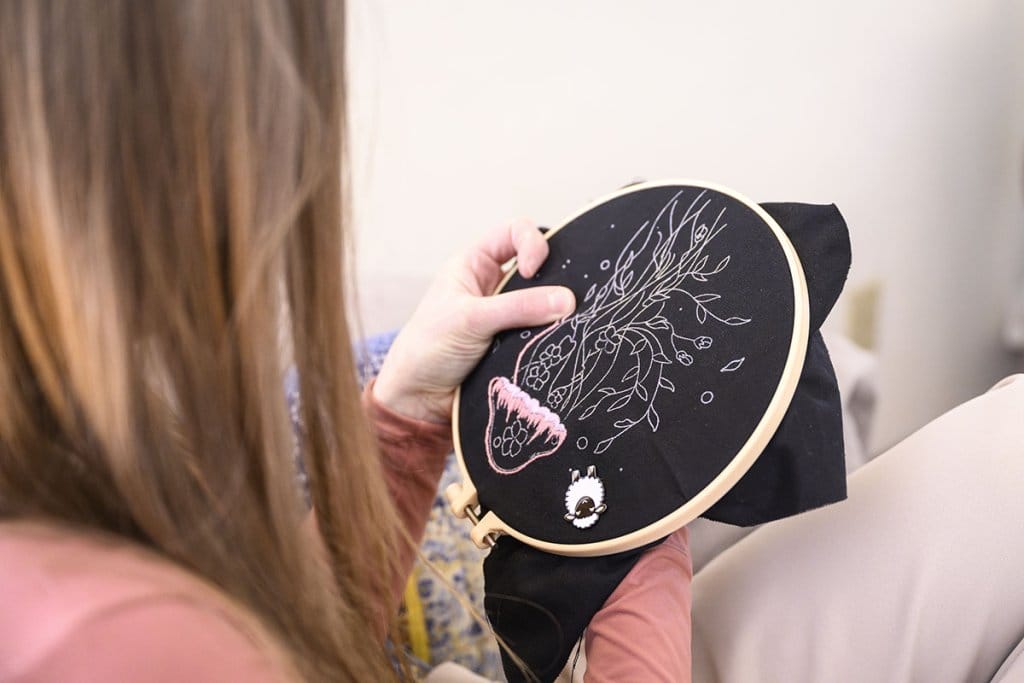 Calarco holds an embroidery hoop with black fabric while working on a floral design with pink and white thread.