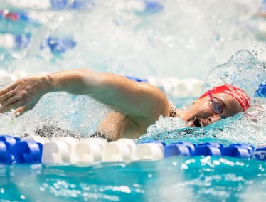 A swimmer wearing a red swim cap is swims freestyle stroke in a pool, with water splashing around and lane lines visible on either side of her.