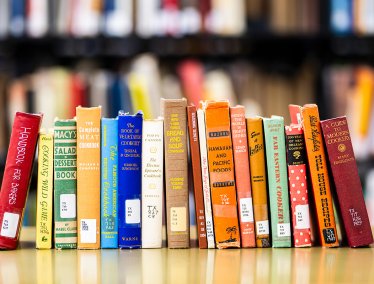 Row of colorful cookbooks standing upright on a table in a library, with blurred bookshelves in the background.