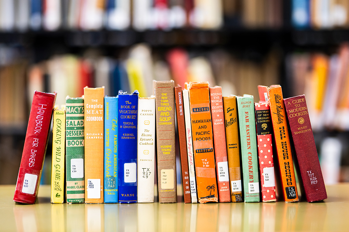 Row of colorful cookbooks standing upright on a table in a library, with blurred bookshelves in the background.