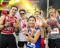 A group of runners with race bibs and medals pose for a photo after a race, with a music stage and colorful lights in the background.