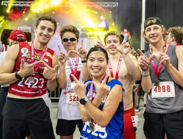 A group of runners with race bibs and medals pose for a photo after a race, with a music stage and colorful lights in the background.