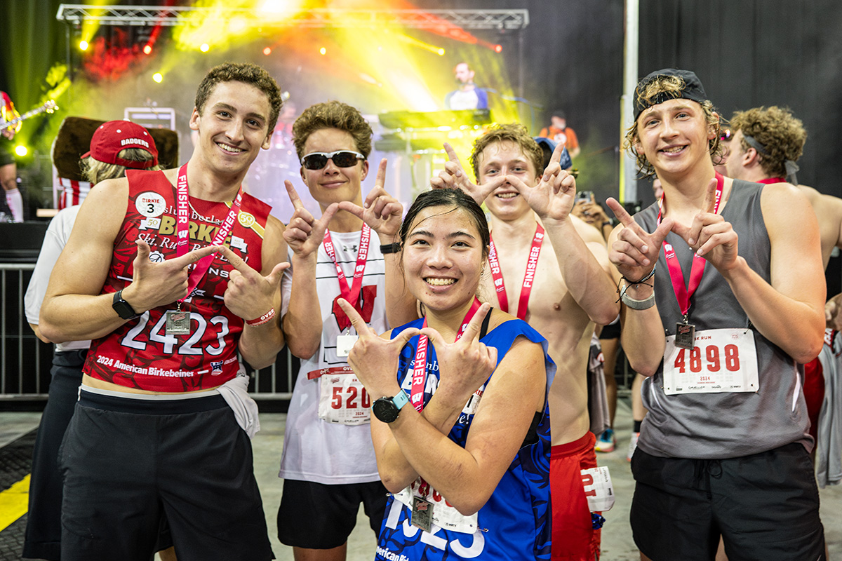 A group of runners with race bibs and medals pose for a photo after a race, with a music stage and colorful lights in the background.