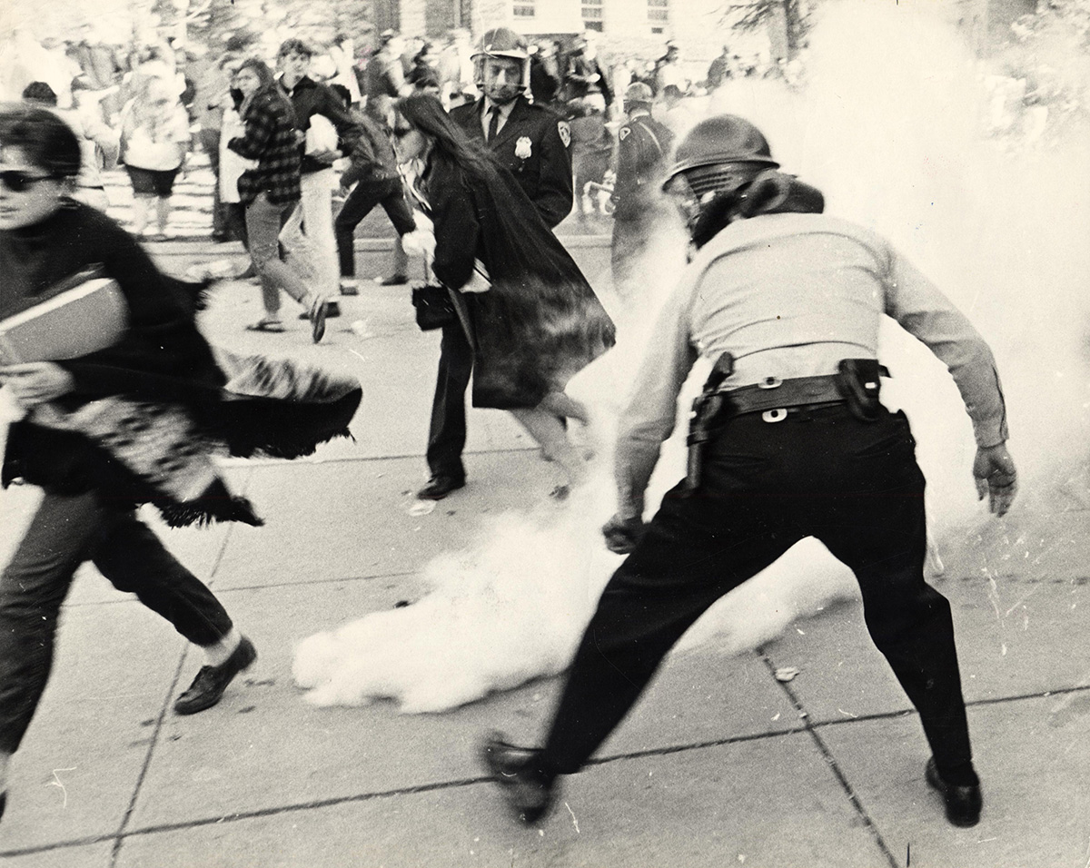 A black and white photograph showing a chaotic scene with people running from a cloud of smoke, while a uniformed police officer in a helmet stands in the foreground.