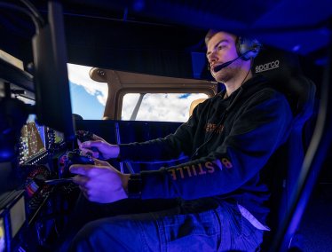 Person seated in a flight simulator cockpit, wearing headphones and using the controls, with screens showing flight instruments and a cloud-filled sky through simulated aircraft windows.