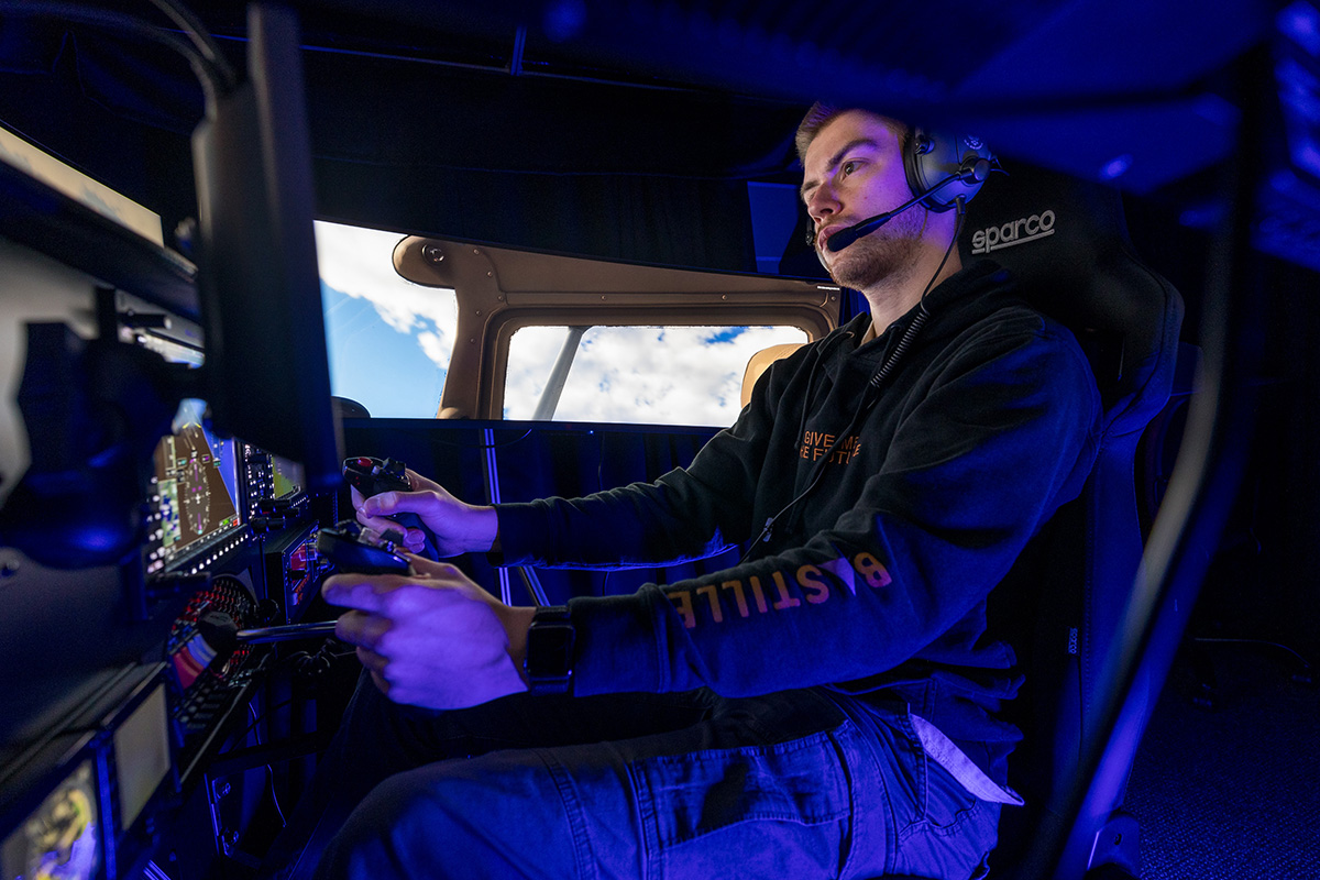 Person seated in a flight simulator cockpit, wearing headphones and using the controls, with screens showing flight instruments and a cloud-filled sky through simulated aircraft windows.