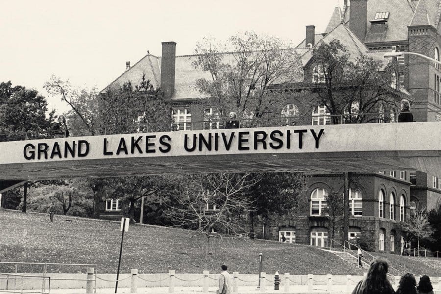 A black and white photograph of the campus bridge that goes over North Park Street labeled 'GRAND LAKES UNIVERSITY,' with science Hall in the background.