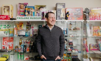 Sean Jacobsohn stands in front of a display case filled with novelty items and memorabilia, beneath a sign that reads 'FAILURE MUSEUM'.
