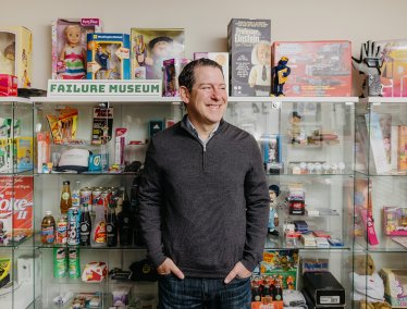 Sean Jacobsohn stands in front of a display case filled with novelty items and memorabilia, beneath a sign that reads 'FAILURE MUSEUM'.