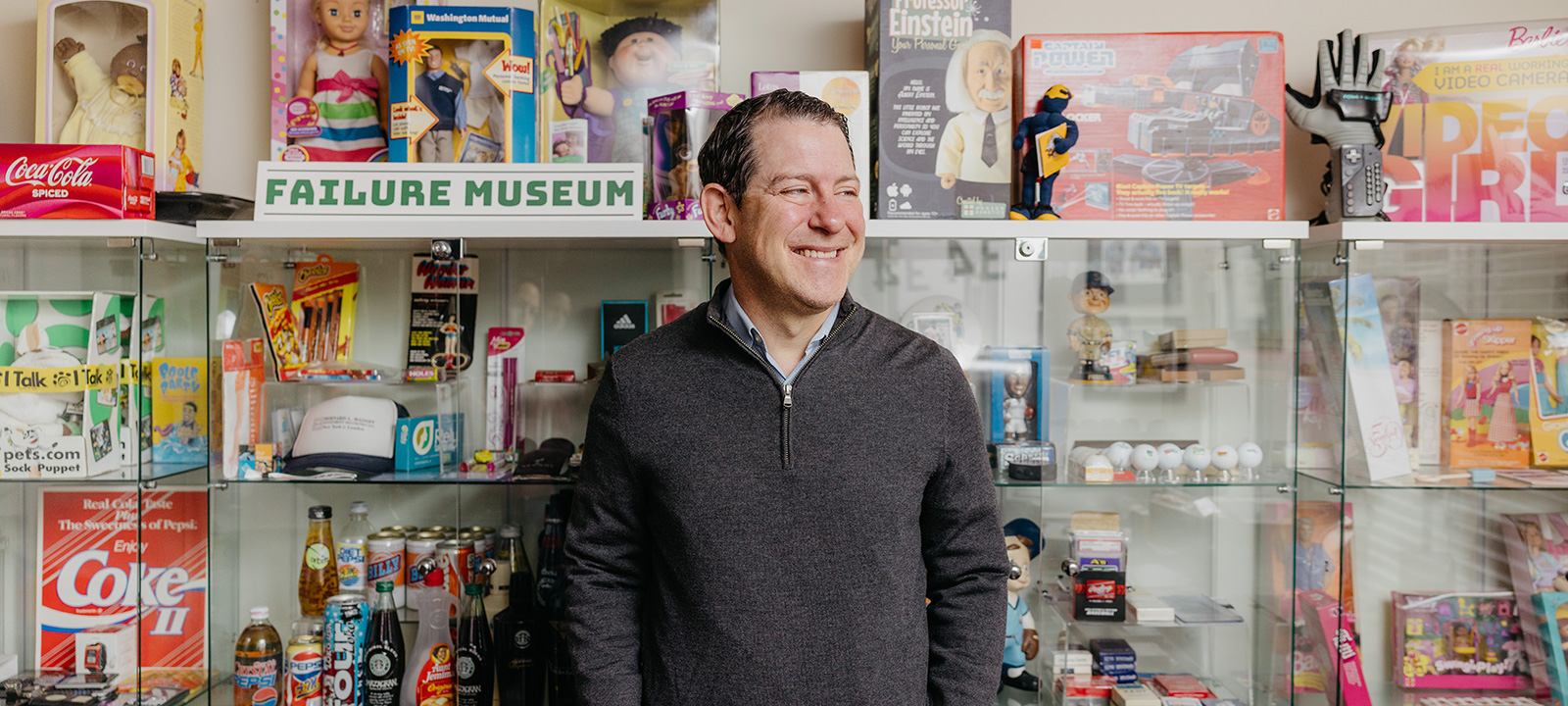 Sean Jacobsohn stands in front of a display case filled with novelty items and memorabilia, beneath a sign that reads 'FAILURE MUSEUM'.