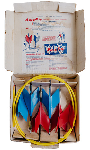 An open box containing a set of Jarts lawn darts, including two blue and two red darts, two yellow plastic rings, and printed instructions on the inside lid.