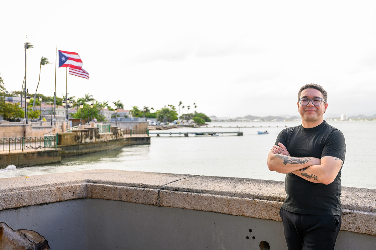 Jorell Meléndez-Badillo standing with arms crossed near a waterfront, with the Puerto Rican flag waving in the background alongside buildings, palm trees, and a dock extending into the water.
