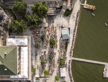 An aerial view of a bustling Memorial Union Terrace with numerous tables and chairs, surrounded by trees and buildings, adjacent to Lake Mendota.
