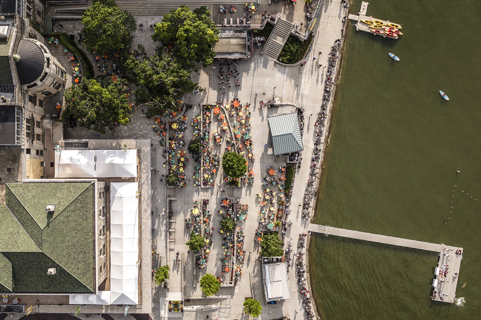 An aerial view of a bustling Memorial Union Terrace with numerous tables and chairs, surrounded by trees and buildings, adjacent to Lake Mendota.
