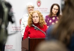 Chancellor Mnookin in a red sweater speaking at a podium with a microphone, in front of two other speakers, and a backdrop featuring the UW-Madison School of Medicine and Public Health logo and name.