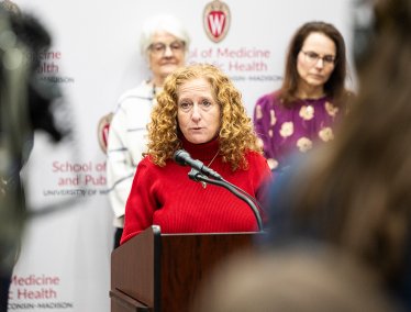 Chancellor Mnookin in a red sweater speaking at a podium with a microphone, in front of two other speakers, and a backdrop featuring the UW-Madison School of Medicine and Public Health logo and name.