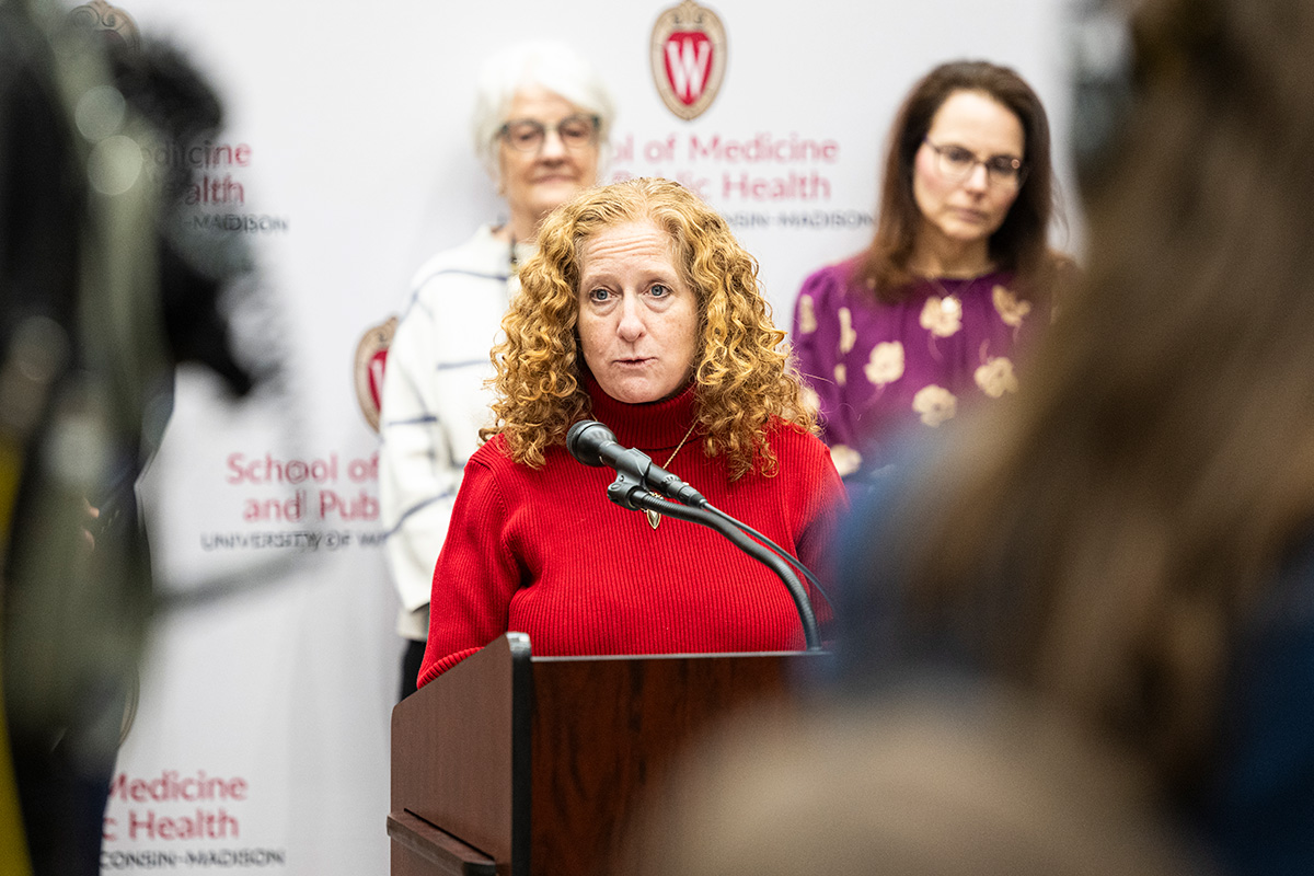 Chancellor Mnookin in a red sweater speaking at a podium with a microphone, in front of two other speakers, and a backdrop featuring the UW-Madison School of Medicine and Public Health logo and name.