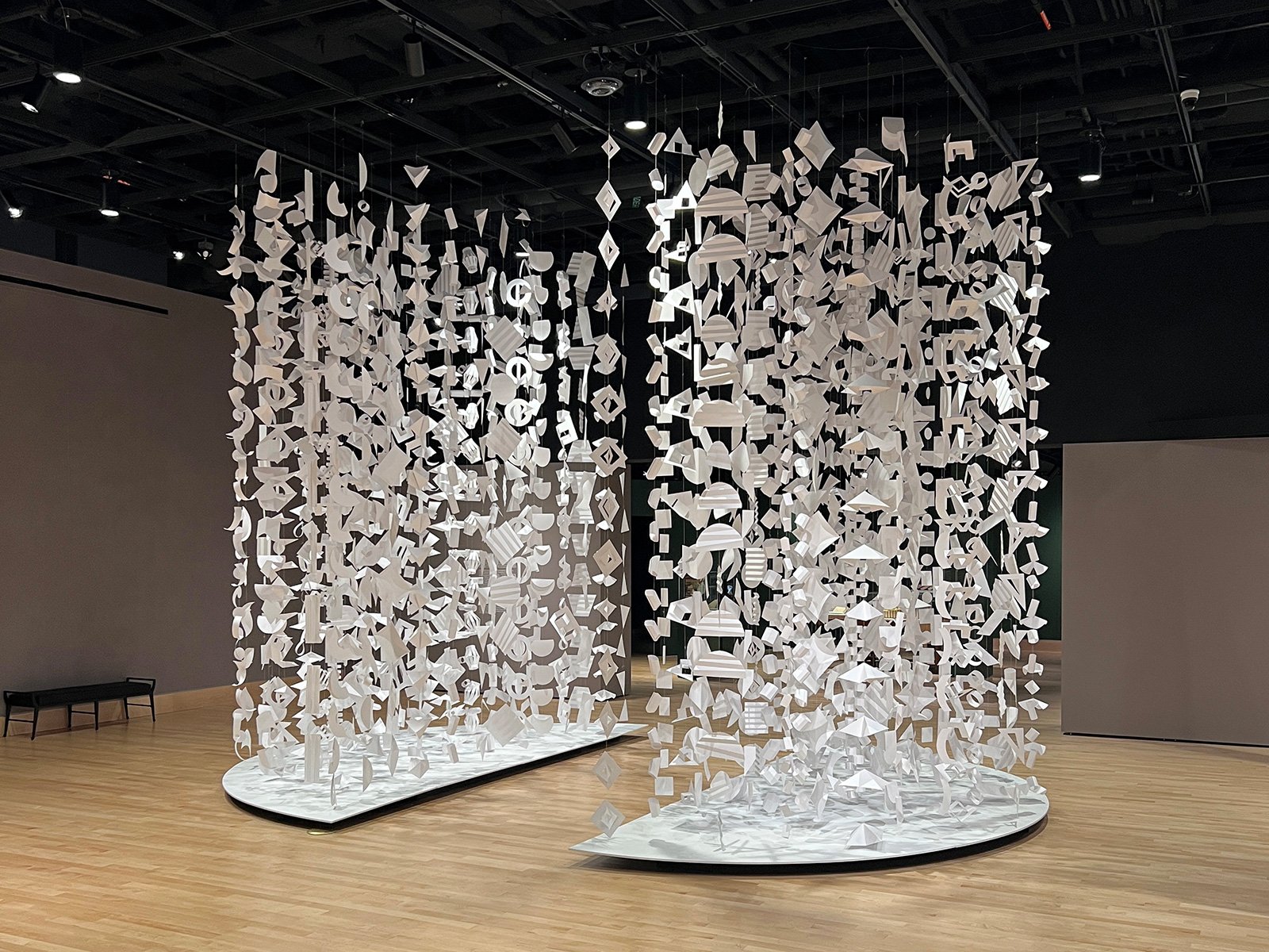 A exhibition piece featuring two large installations composed of numerous white paper shapes suspended from the ceiling in a gallery space with wooden floors and dark walls.