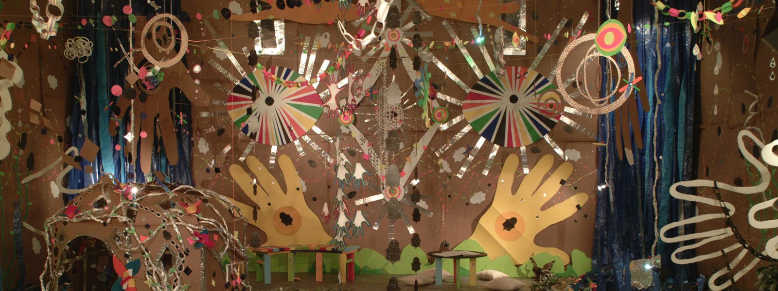 A colorful and whimsical art installation featuring large hand cutouts, vibrant circular patterns, hanging decorations, a small bridge with rainbow stripes, and various abstract elements made from paper, cardboard and foil.