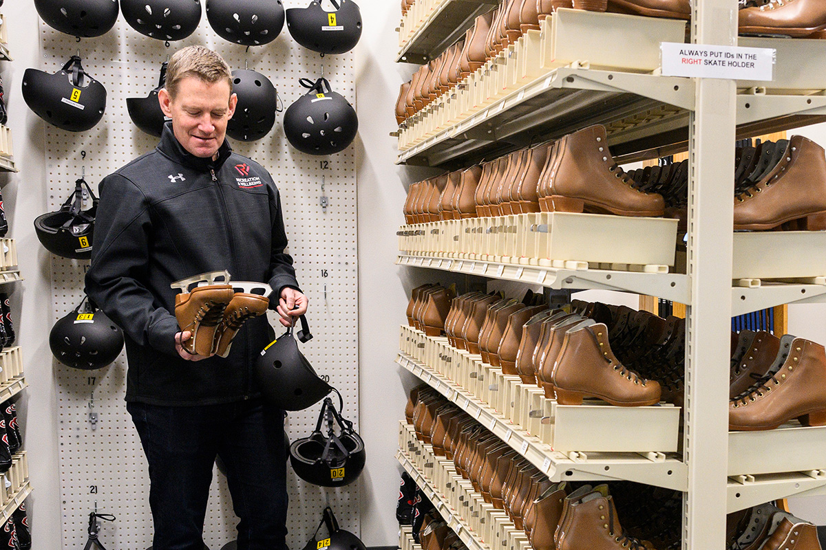 Bill Riggins holding brown ice skates and a black helmet in an area lined with shelves full of helmets and ice skates.