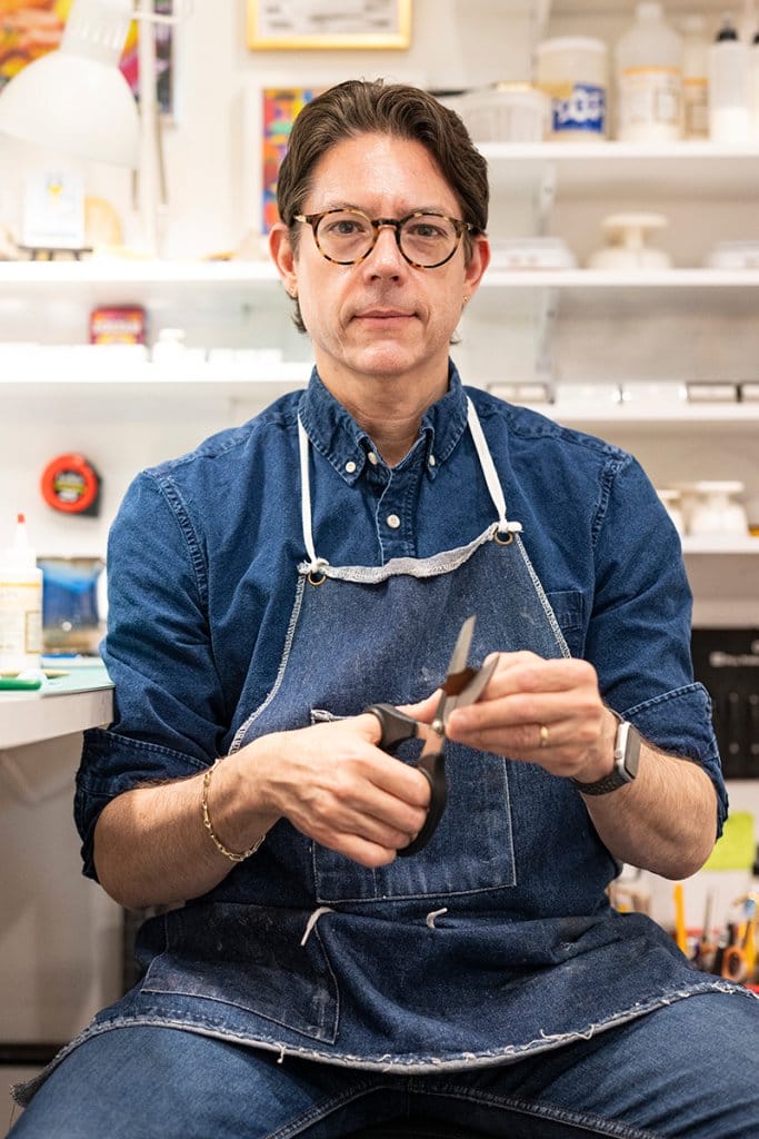 Velliquette sitting in his workshop in a blue denim shirt and apron, holding a pair of scissors with both hands.