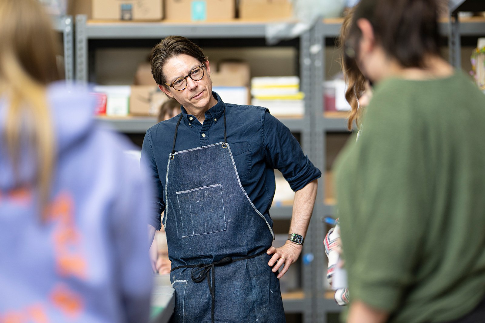 Michael Velliquette in a blue shirt and denim apron stands with hands on hips in a workshop, surrounded by three other individuals.