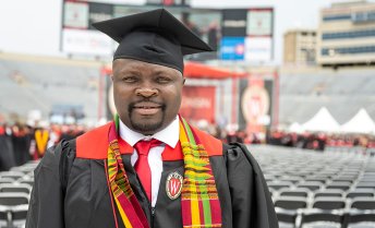 A graduate wearing a cap and gown, adorned with a red and yellow stole, stands in Camp Randall Stadium with other graduates in the background.