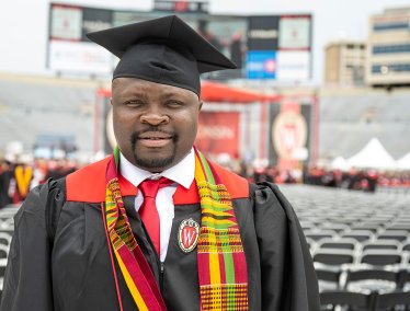 A graduate wearing a cap and gown, adorned with a red and yellow stole, stands in Camp Randall Stadium with other graduates in the background.
