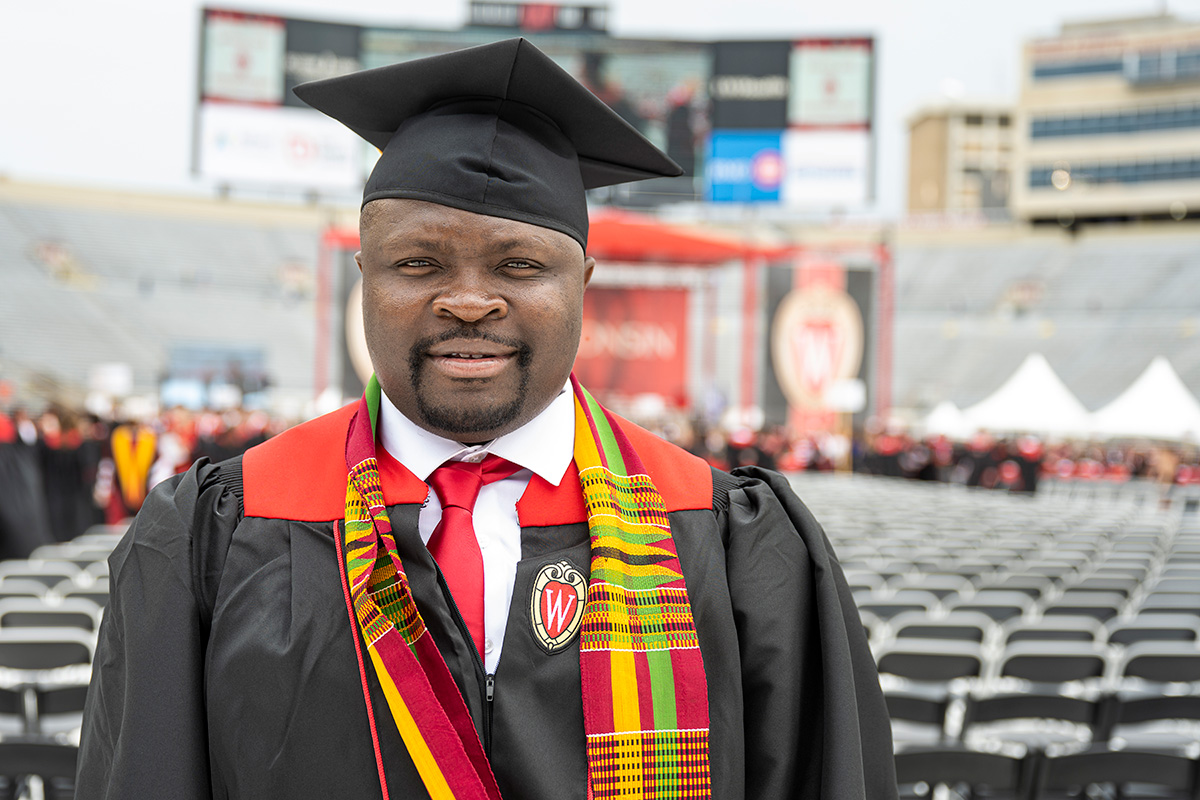 A graduate wearing a cap and gown, adorned with a red and yellow stole, stands in Camp Randall Stadium with other graduates in the background.
