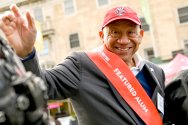 Elzie Higgenbottom smiling and waving, wearing a red Badger cap and red sash