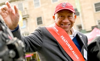 Elzie Higgenbottom smiling and waving, wearing a red Badger cap and red sash