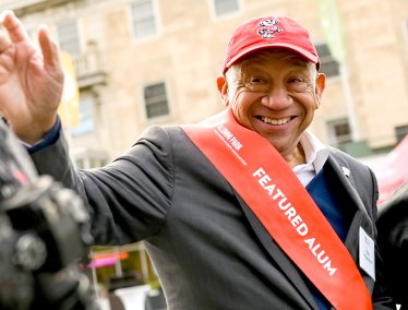Elzie Higgenbottom smiling and waving, wearing a red Badger cap and red sash