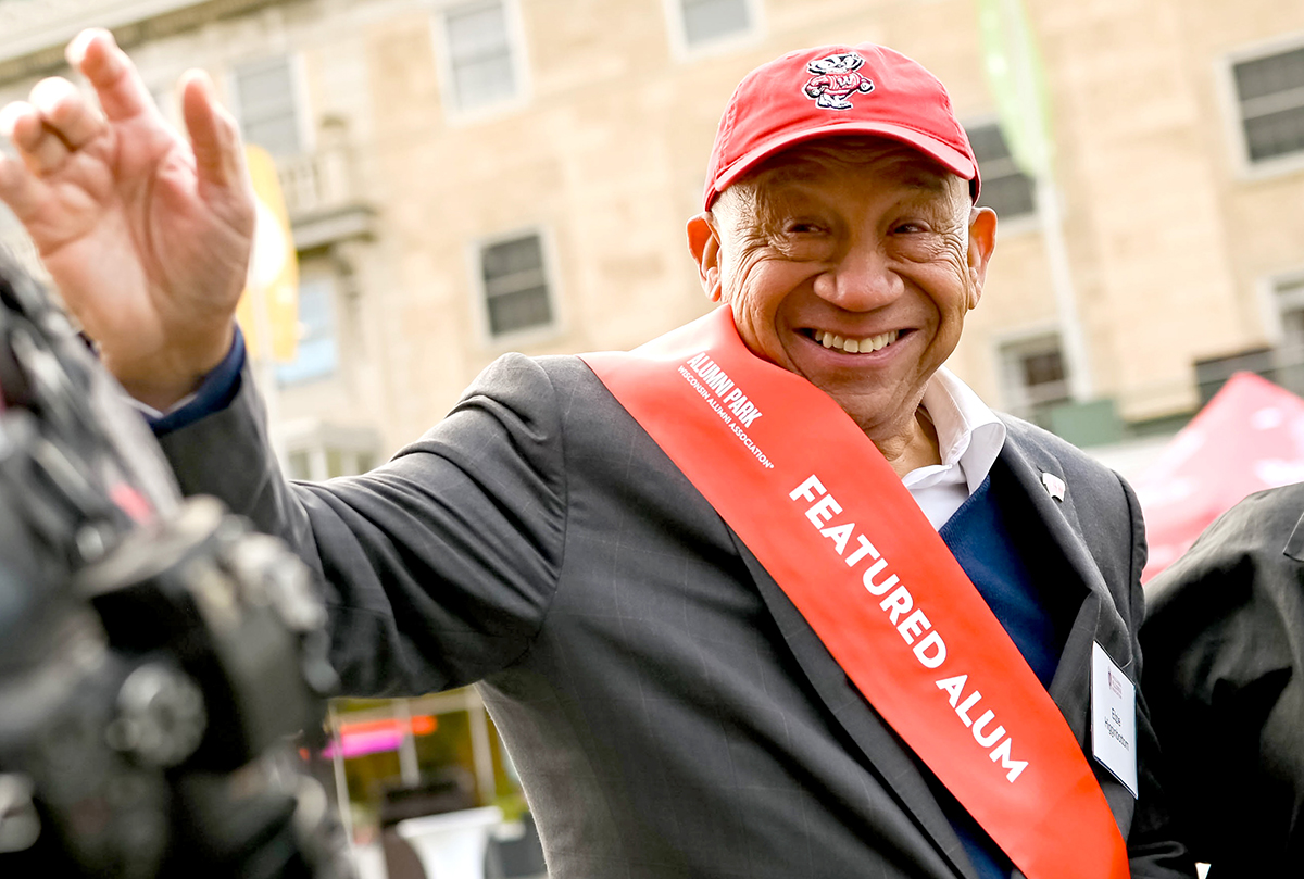 Elzie Higgenbottom smiling and waving, wearing a red Badger cap and red sash