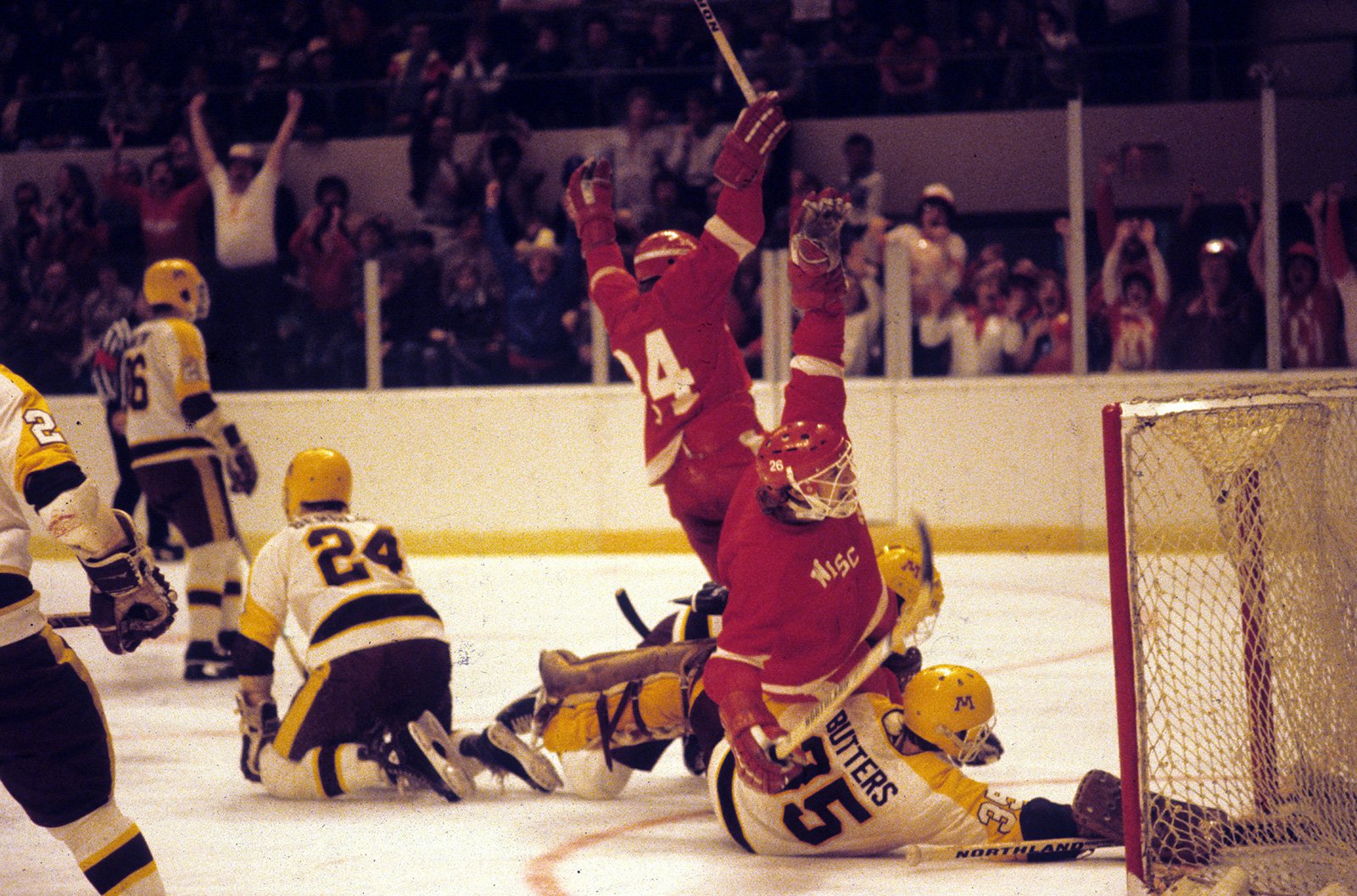 During an ice hockey game, two Badger players in red jerseys raise their arms in celebration near the goal, while a Gopher lies on the ice and another kneels nearby, with a cheering crowd in the background.