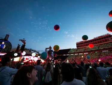 A lively outdoor concert scene shows a cheering crowd beneath a clear evening sky, with colorful balloons floating and a brightly lit stage featuring Chris Martin from Coldplay on a large screen.