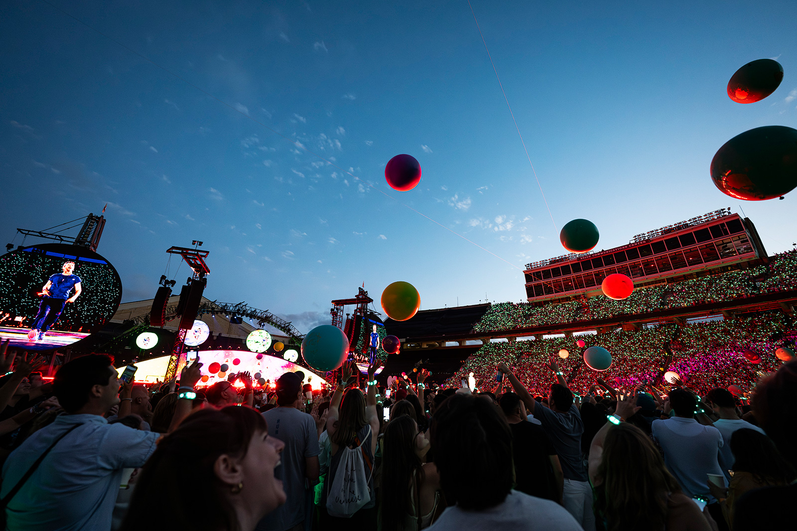 A lively outdoor concert scene shows a cheering crowd beneath a clear evening sky, with colorful balloons floating and a brightly lit stage featuring Chris Martin from Coldplay on a large screen.