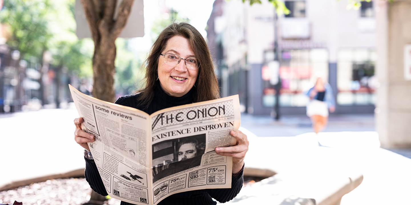 Christine Wenc sits outside reading a newspaper titled 'The Onion,' with the bold headline 'Existence Disproven,' surrounded by playful sections and ads.