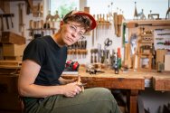 Katie Hudnall sits in a well-equipped woodworking workshop, carving a small piece of wood.
