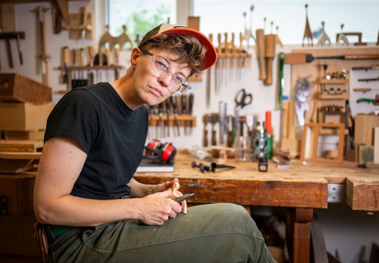 Katie Hudnall sits in a well-equipped woodworking workshop, carving a small piece of wood.