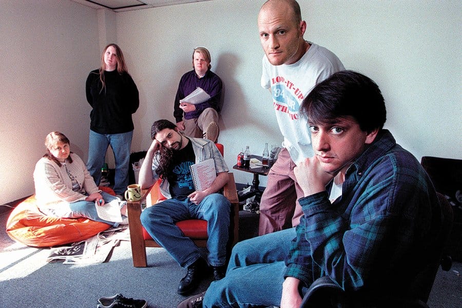 Six people, some seated, some standing, pose in a room with casual furniture including a beanbag. All of them look intently, not smiling, at the camera.