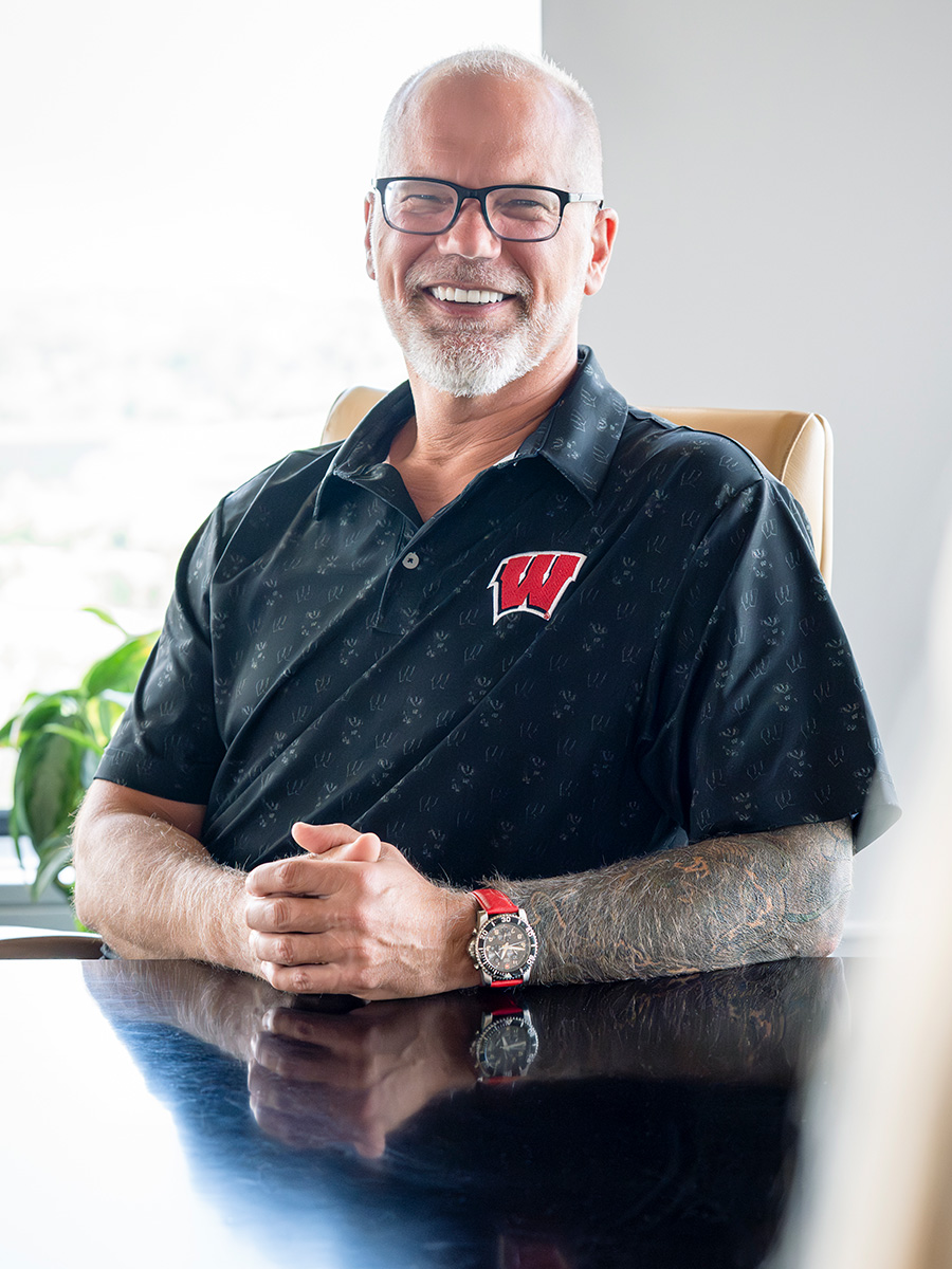 Eric Iverson, wearing a dark polo shirt with a red 'W' logo sits at a boardroom table, revealing a tattooed arm and red-banded wristwatch.