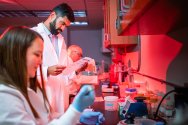 In a red-lit laboratory, three people in white lab coats conduct research—one reads a document, another uses a pipette, and the lab counter is filled with scientific equipment.