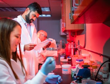 In a red-lit laboratory, three people in white lab coats conduct research—one reads a document, another uses a pipette, and the lab counter is filled with scientific equipment.