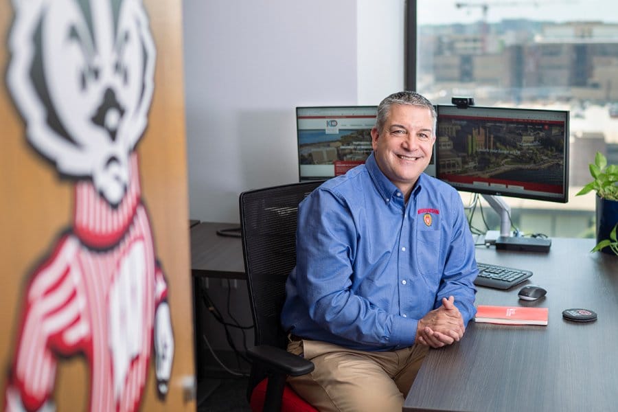 Greg Keenan, wearing a blue button-up 'Wisconsin' shirt sits at an office desk with dual monitors displaying the UW-Madison website, surrounded by items including a large Bucky Badger cutout on a door.