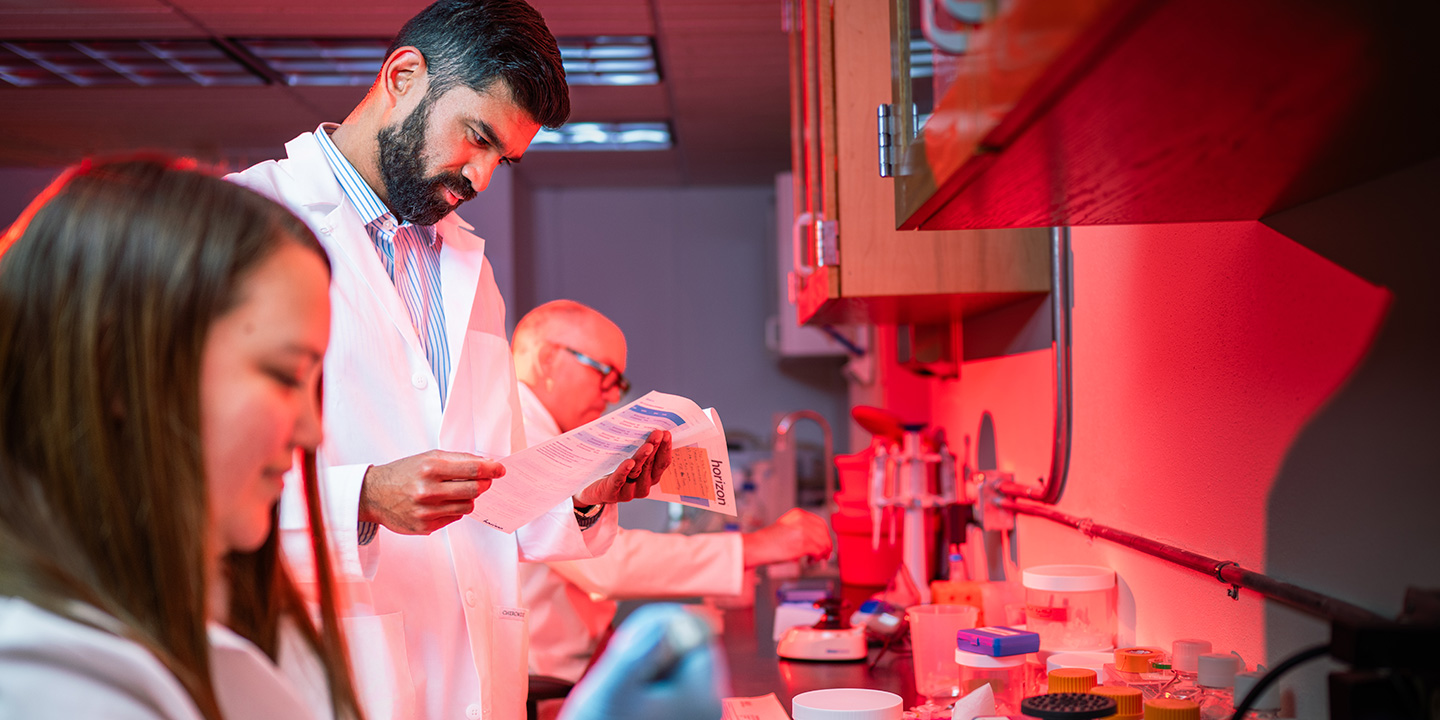 In a red-lit laboratory, three people in white lab coats conduct research—one reads a document, another uses a pipette, and the lab counter is filled with scientific equipment.