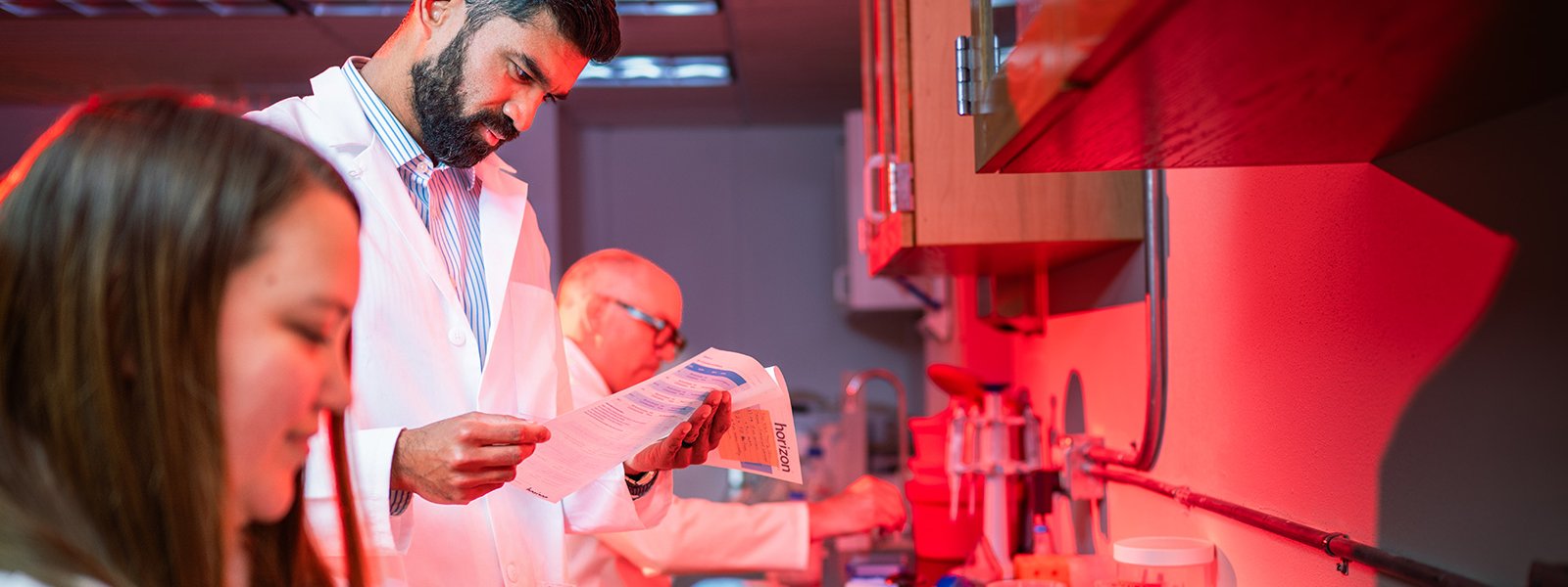 In a red-lit laboratory, Faraz Choudhury and two other people in white lab coats conduct research --Choudhury reads a document, another uses a pipette, and the lab counter is filled with scientific equipment.