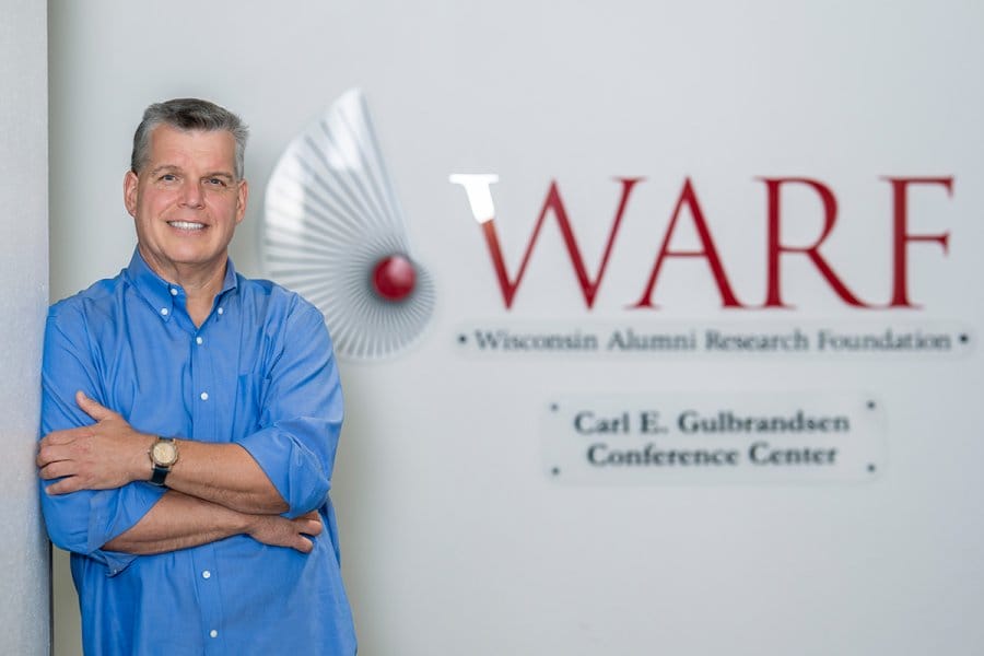 Mike Partsch, wearing a blue button-up shirt, stands with arms crossed beside a white wall featuring the WARF logo.