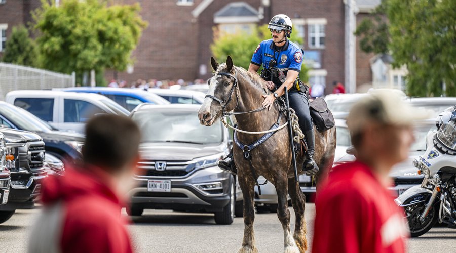 A mounted police officer rides a gray horse through a parking lot with cars and buildings in the background.
